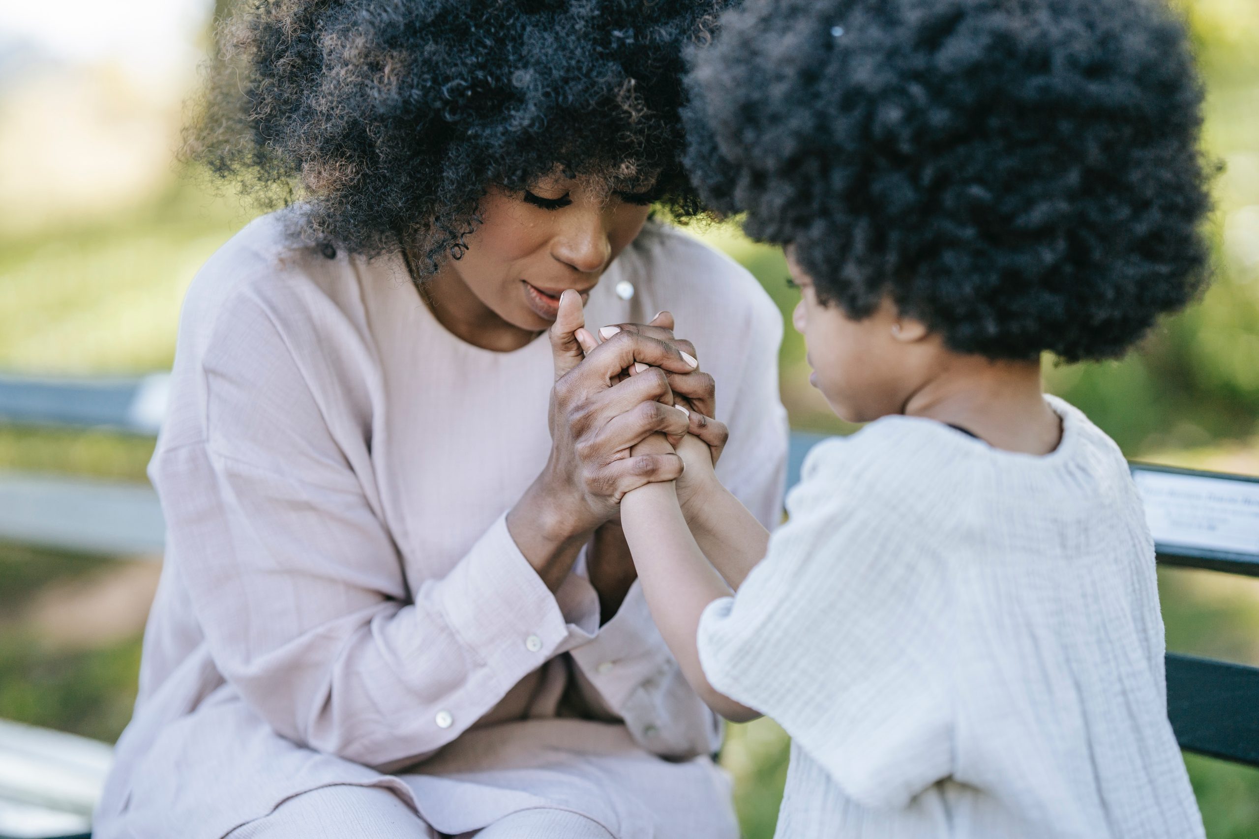Close up of mom holding her child's hands and praying with her while sitting on a bench outside