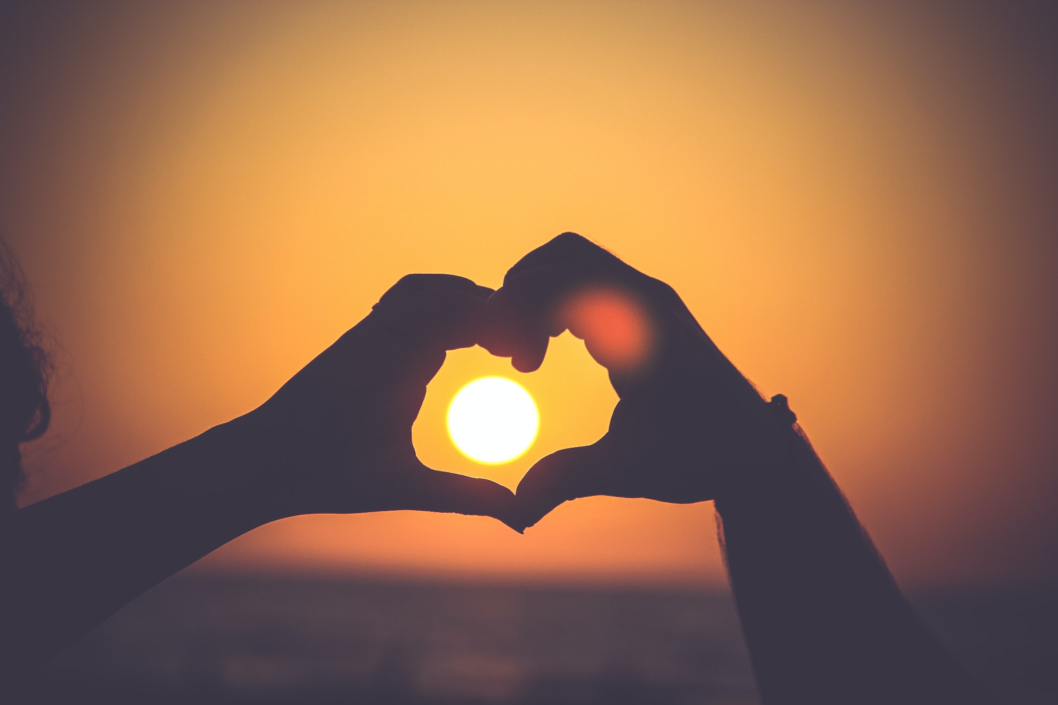 Close up of hands forming a heart, framing it over the setting sun in the background